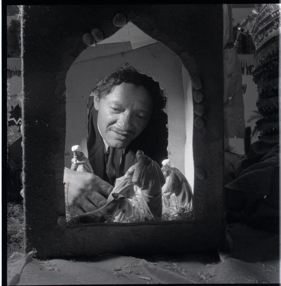 Hombre frente a pesebre. Esta fotografía composicionalmente muy destacada, juega con los varios planos creados por lo que parece ser la ventana de cartón de un pesebre. El marco creado por el fotógrafo enfatiza en el protagonismo del hombre que construye su pesebre.