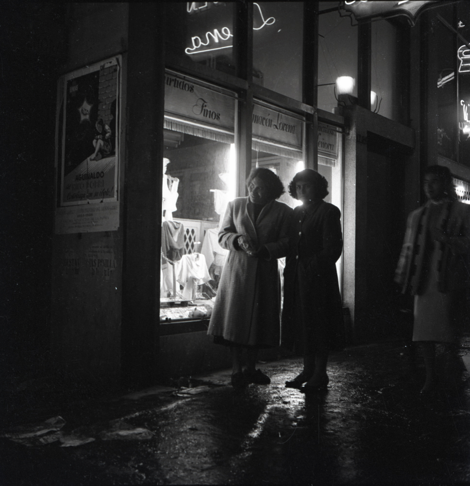 Tomada durante la noche de año nuevo bogotana en 1956, esta fotografía captura a dos mujeres vestidas de abrigo recorriendo la ciudad. Caicedo aprovecha la luz de las vitrinas para iluminar a las caminantes, capturando no solo las retratadas, sino también la actividad comercial de una Bogotá que se presenta como moderna y cosmopolita. El cartel en el lado izquierdo de la fotografía así como el letrero en luz neón que se vislumbra en el fondo, hablan de una inminente modernidad en la Bogotá de mediados del siglo XX.