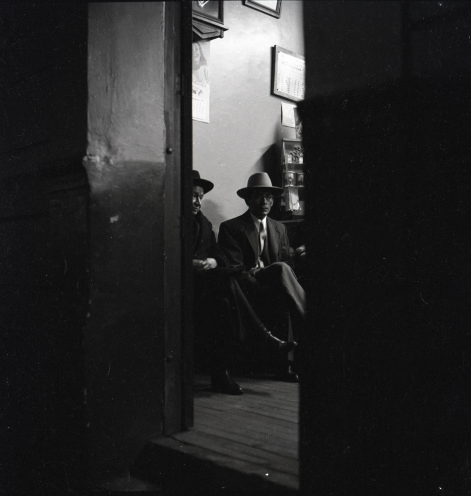 En esta fotografía Caicedo captura desde el exterior de la calle, una escena en el interior de una tienda de una ciudad. La escena principal está enmarcada por la puerta de la edificación y en ella se ven dos personas sentadas: un hombre de traje, corbata y sombrero que se contrapone con el perfil de una mujer vestida con ropa más tradicional. La oscuridad de la noche contrasta con la luz interior de la tienda creando un ambiente de misterio en una escena de la vida cotidiana que el fotógrafo captura como si fuera un voyeur.