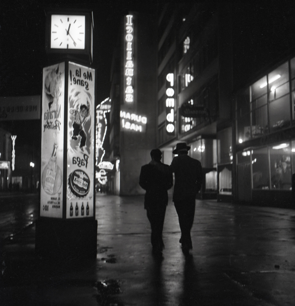 Dos caminantes aparecen de espaldas en el centro de la imagen tomada en Bogotá a las 11:36 pm como lo indica el reloj a la derecha de la imagen. La fotografía remite al ambiente de una película de cine negro, en donde el uso de sombras y claroscuros es prominente y ayuda a crear dramatismo en la escena. La oscuridad de la noche contrasta con la luz que emanan los letreros de publicidad y que nos hablan de una ciudad netamente moderna. 