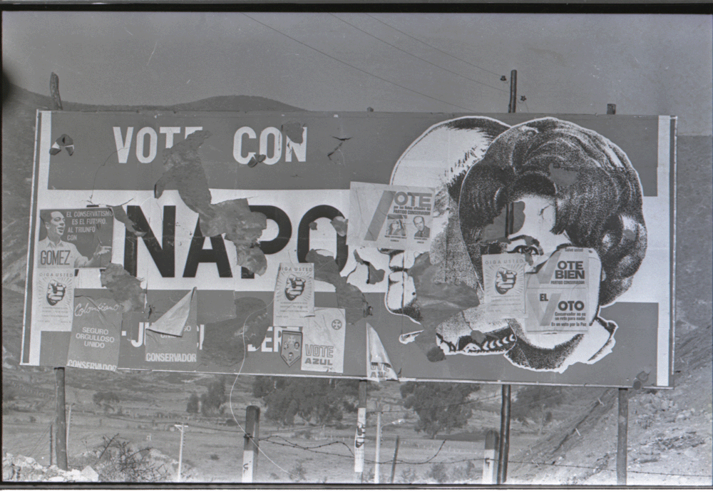 Foto de una valla publicitaria de María Eugenia Rojas del partido político ANAPO (Alianza Nacional Popular) durante las elecciones presidenciales de 1974, las cuales ganó Alfonso López Michelsen. En la valla, María Eugenia Rojas aparece delante de la figura de su papá, el general Gustavo Rojas Pinilla quien, para el momento de la foto y de la valla, había recuperado sus derechos políticos, después del golpe de estado que recibió en 1957. La valla está cubierta por carteles, uno de ellos de Álvaro Gómez Hurtado, del partido Conservador, quien también fue contrincante en dichas elecciones. (Sobre cuestiones generales de la ANAPO, ver Marco Palacios y Frank Safford, Colombia. País fragmentado, sociedad dividida, Bogotá: Editorial Norma, 2006, 605-610.)